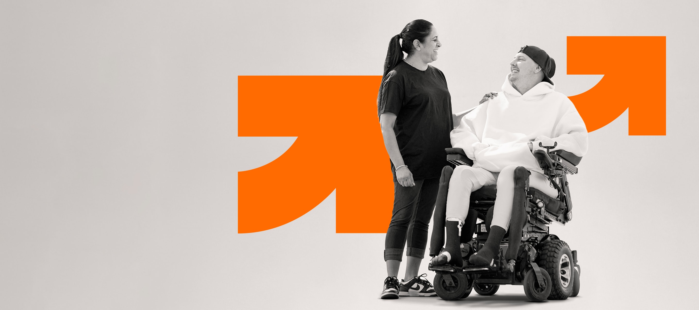 A carer with her hand on the shoulder of a smiling young man in a power wheelchair, the orange Drake arrow behind them