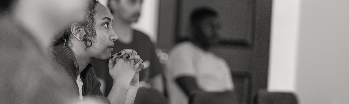 A young woman listening intently, hands clasped under her chin, in a workshop